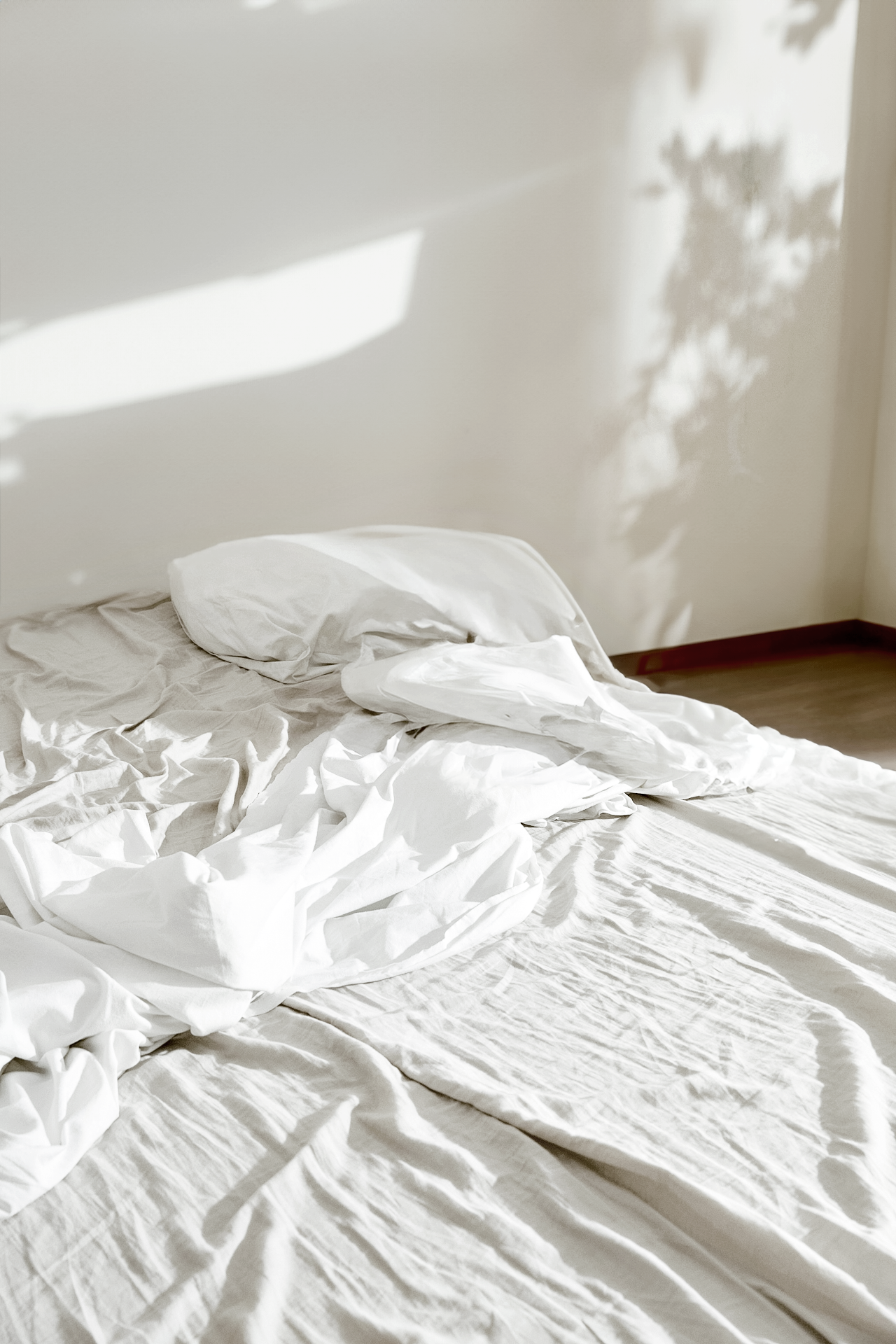 Unmade bed with white sheets and pillow in a room with sunlight casting shadows on the wall.