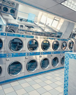 A laundromat with multiple stacked front-loading washing machines and dryers, with some windows allowing natural light in.