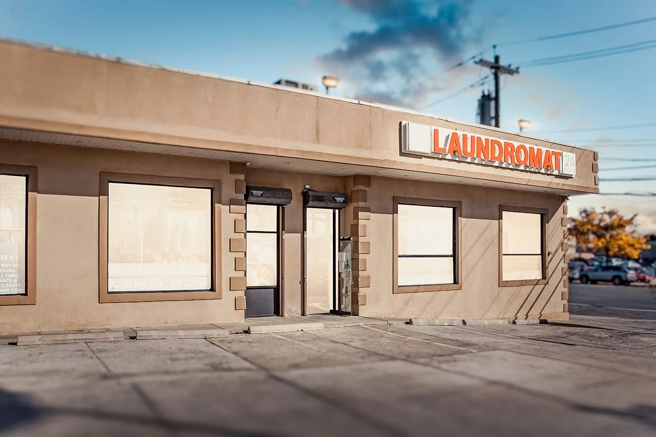Empty laundromat storefront with a sign reading 'LAUNDROMAT' in orange letters, large front windows, and a parking lot in the background, during late afternoon with clear sky and some clouds.