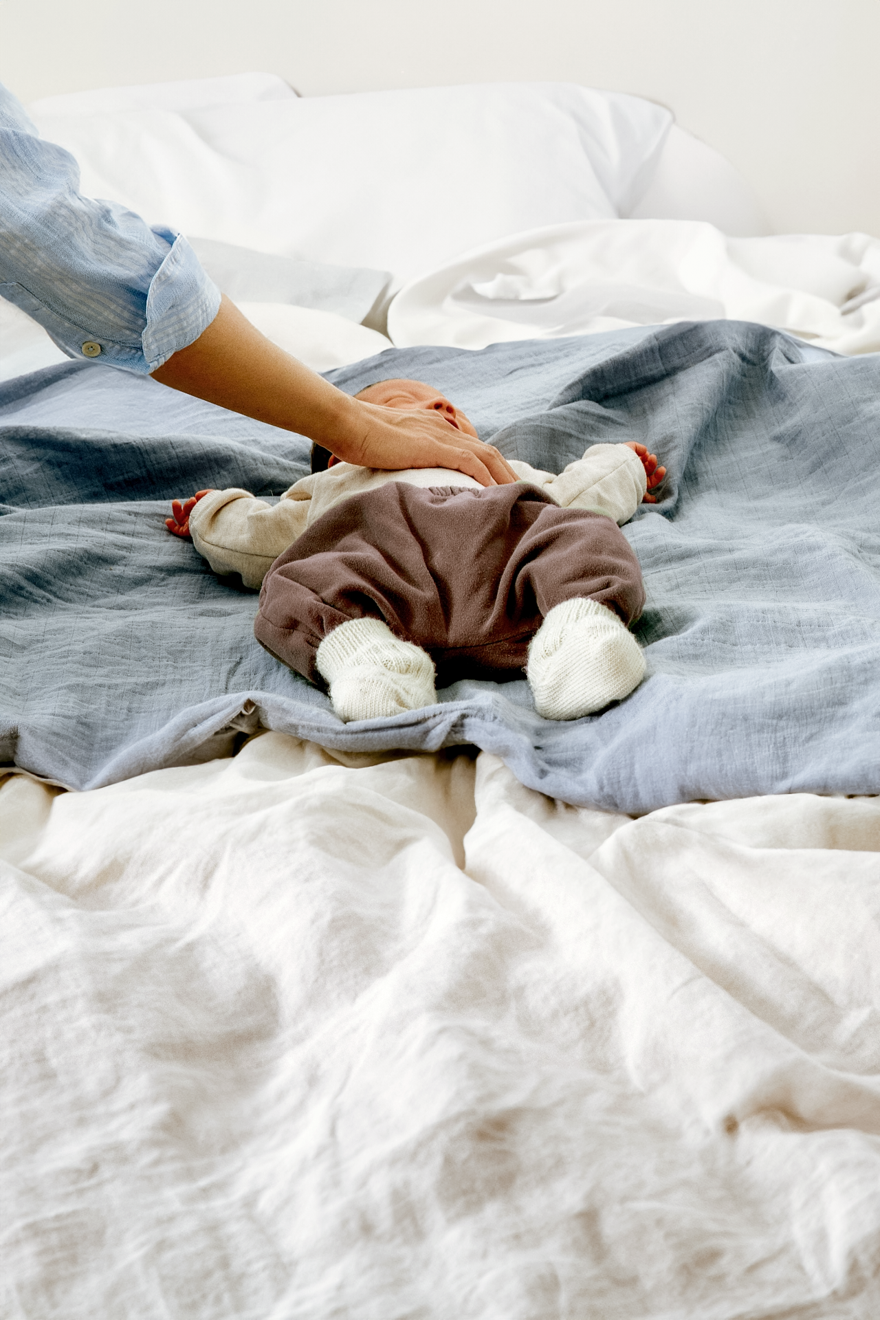 A baby lying face down on a bed, with a hand gently touching the baby's back. The bed has layered white and gray blankets and sheets, and the baby is wearing a cream sweater, brown pants, and white socks.