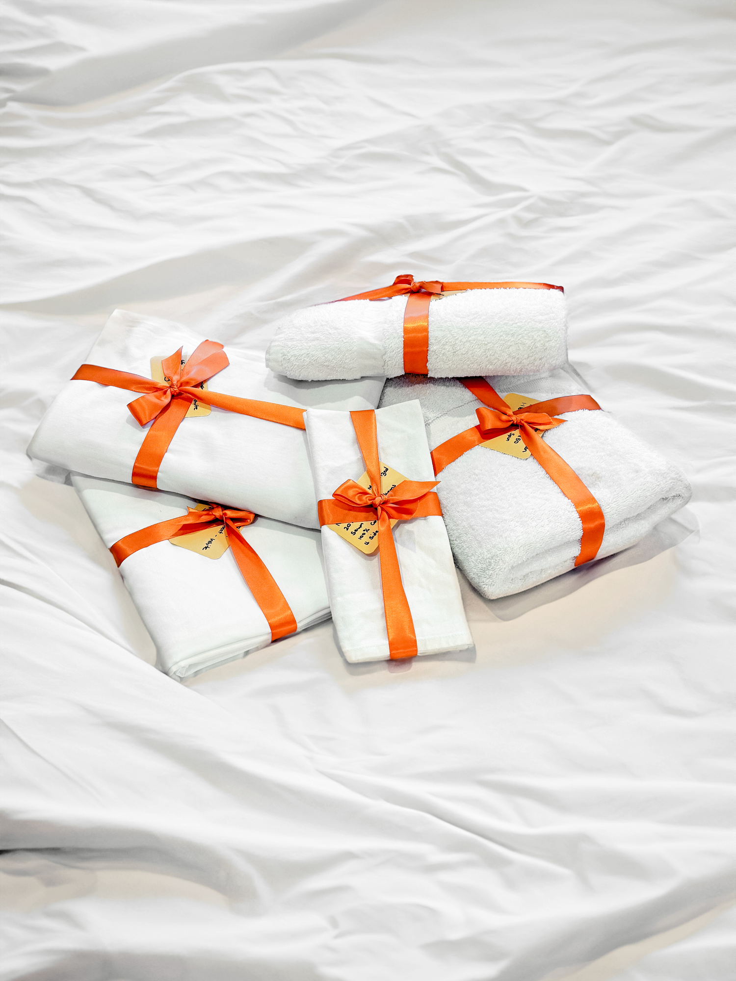 Five white towels and linens, each wrapped with orange ribbons and tags, arranged on a white, crinkled fabric background.