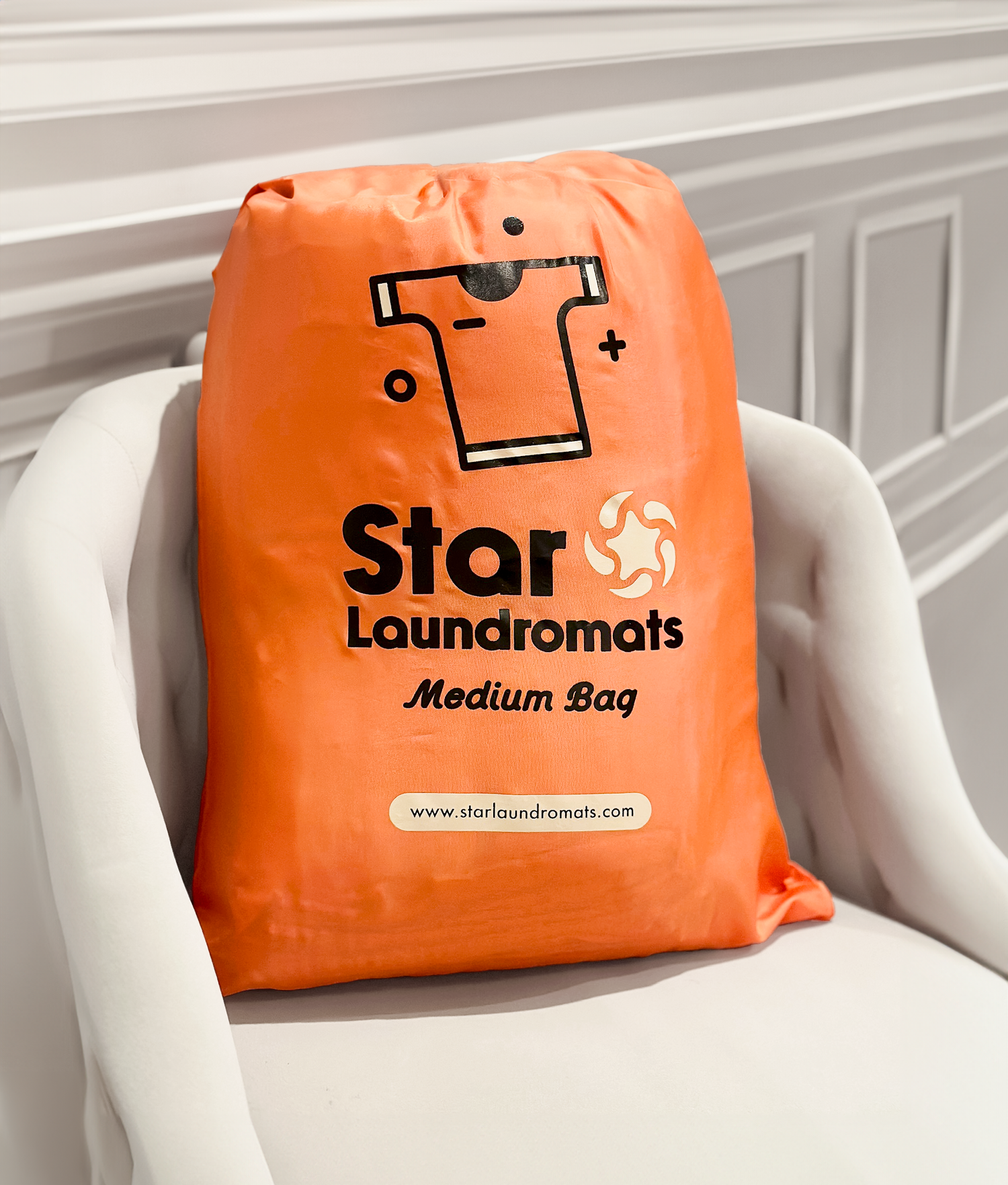 Orange medium-sized laundry bag from star laundromats, placed on a white armchair in a room with white paneling.
