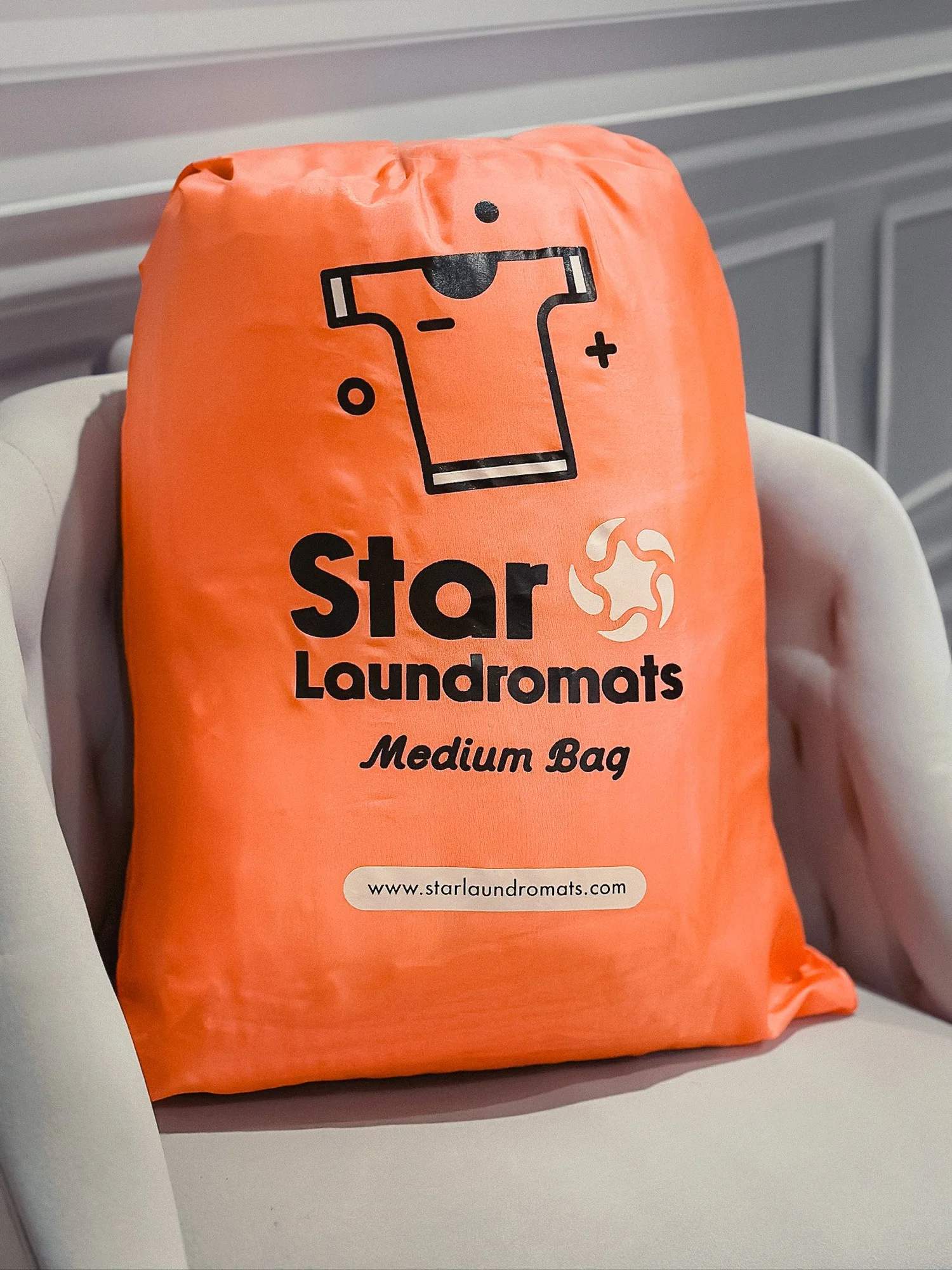 Orange medium-sized laundry bag labeled 'Star Laundromats' with a detergent icon and a website URL, placed on a white chair.