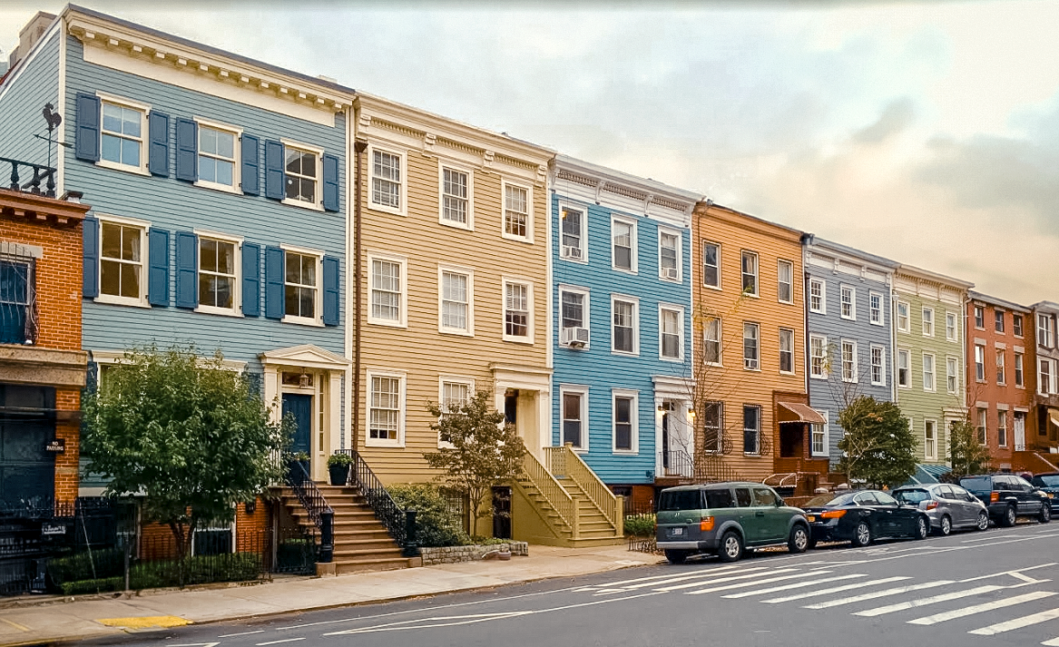 Colorful row of townhouses in pastel shades, with parked cars in front, trees lining the sidewalk, and a cloudy sky above.