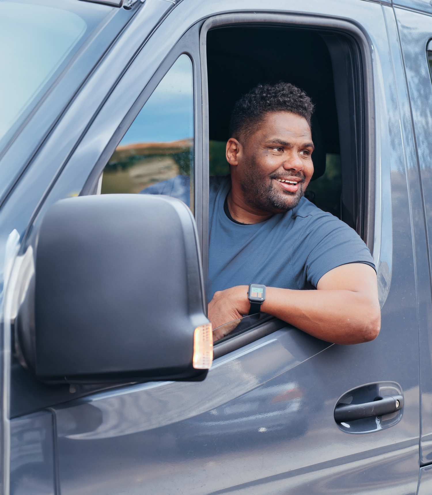 A man with dark hair and a beard smiling while leaning out of the window of a dark gray pickup truck, wearing a gray T-shirt and a smartwatch.
